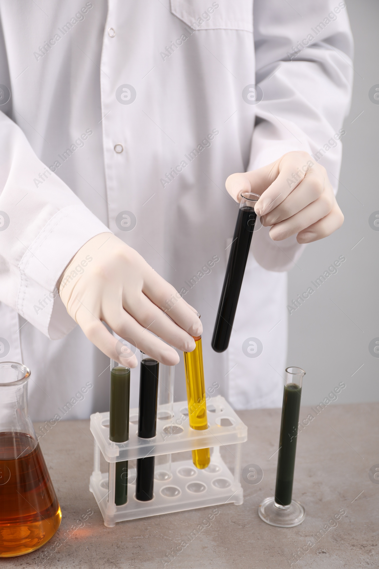 Photo of Woman taking test tubes with different types of crude oil from rack at grey table against light background, closeup