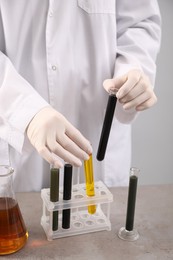 Woman taking test tubes with different types of crude oil from rack at grey table against light background, closeup