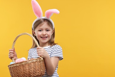 Photo of Easter celebration. Cute girl with bunny ears holding basket of painted eggs on orange background, space for text