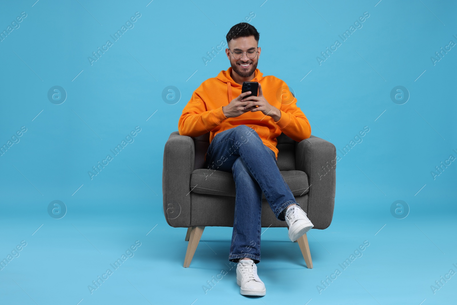 Photo of Happy young man using smartphone on armchair against light blue background