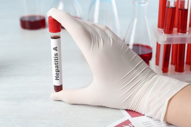 Scientist holding tube with blood sample and label Hepatitis A at white wooden table, closeup