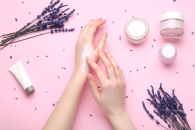 Woman applying hand cream and lavender flowers on pink background, top view