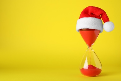 Photo of Hourglass and Santa hat on yellow background, space for text. Christmas countdown