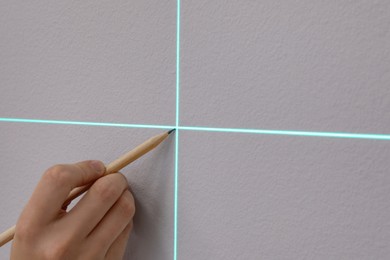 Woman using cross line laser level and pencil for accurate measurement on grey wall, closeup