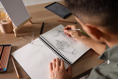 Photo of Man drawing in sketchbook with pencil at wooden table, closeup