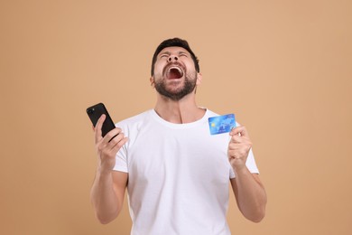 Photo of Emotional man with credit card and smartphone on beige background. Be careful - fraud