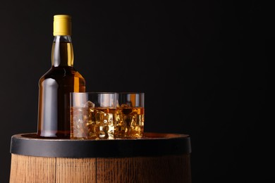 Photo of Whiskey with ice cubes in glasses and bottle on wooden barrel against black background, space for text