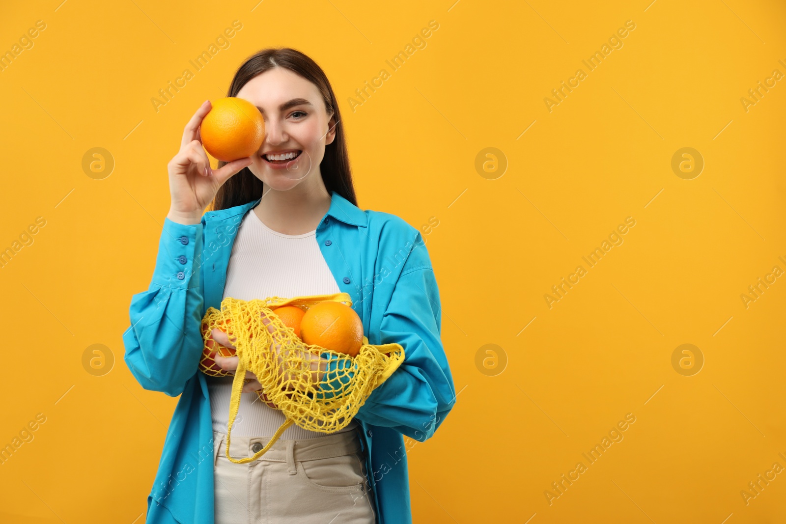 Photo of Woman holding string bag of fresh oranges and covering eye with fruit on orange background, space for text