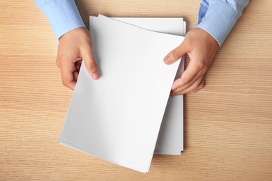 Man holding blank paper sheets for brochure at wooden table, top view. Mock up