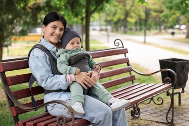 Mother holding her child in sling (baby carrier) on bench in park. Space for text