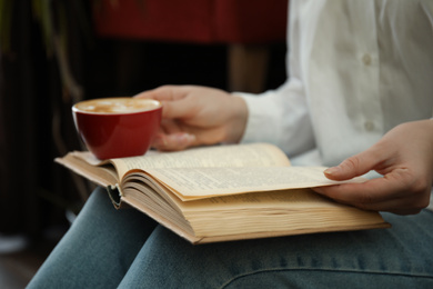 Woman with cup of coffee reading book indoors, closeup