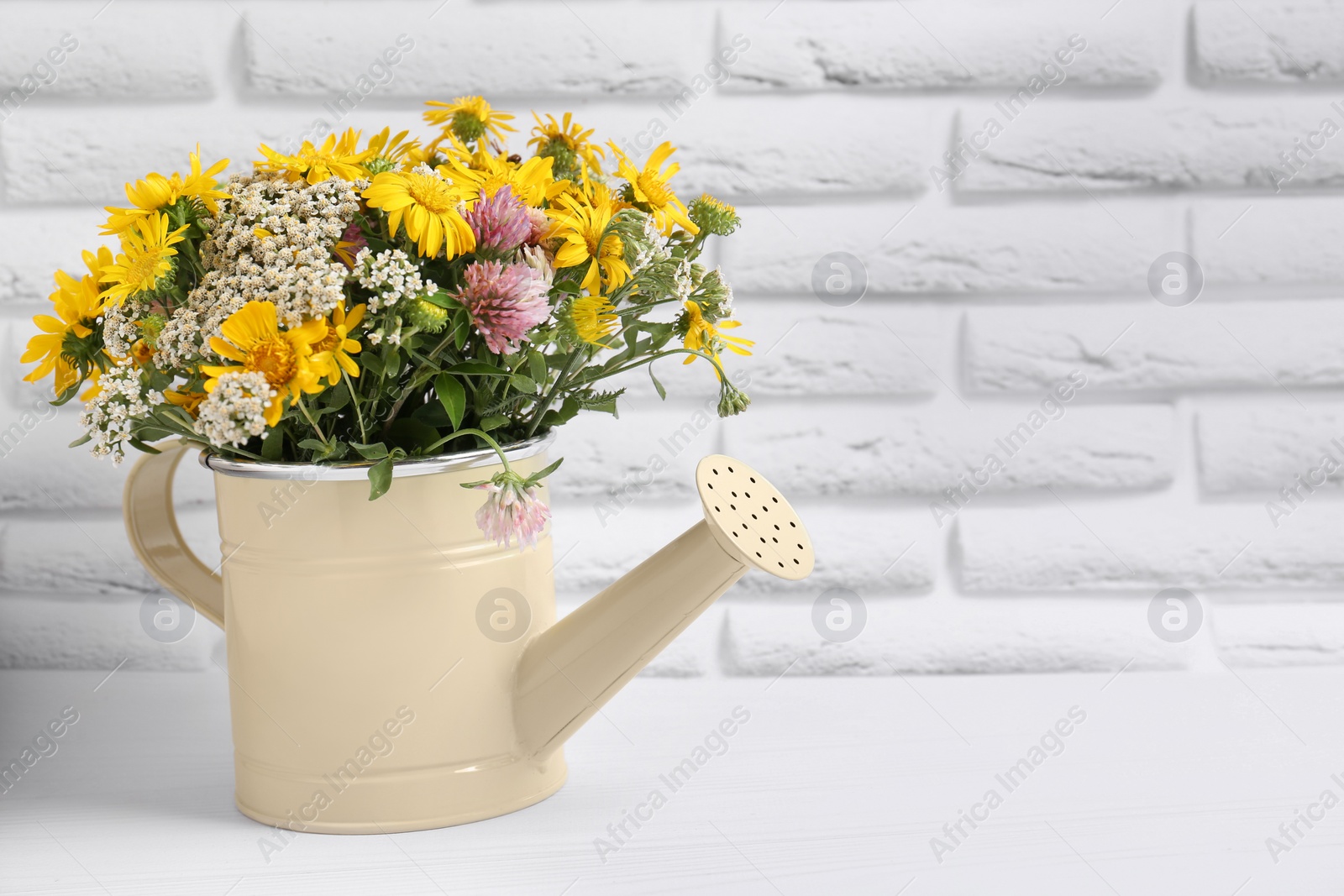 Photo of Beautiful bouquet of bright wildflowers in watering can on white wooden table near brick wall, space for text