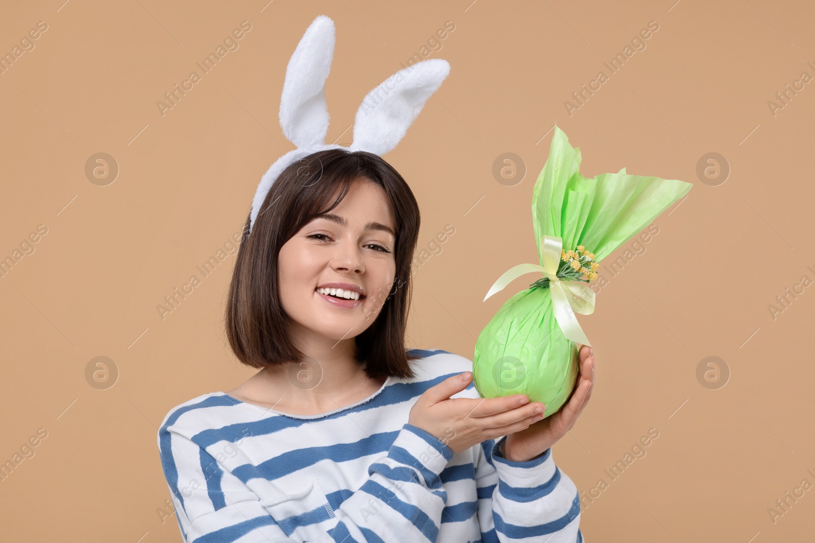 Photo of Easter celebration. Happy woman with bunny ears and wrapped egg on beige background