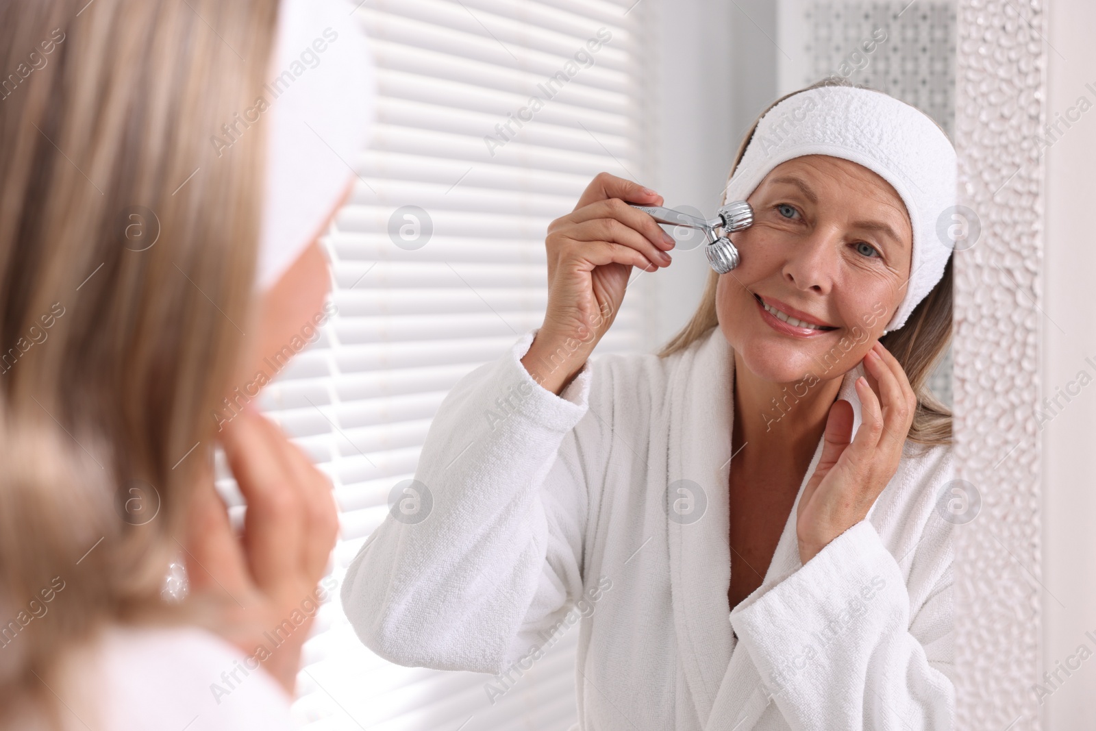 Photo of Woman massaging her face with metal roller near mirror in bathroom