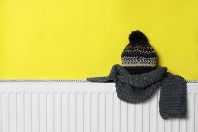 Knitted hat and scarf on heating radiator near yellow wall, space for text