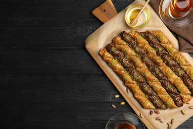 Photo of Delicious sweet baklava with honey and hot tea on black wooden table, flat lay. Space for text