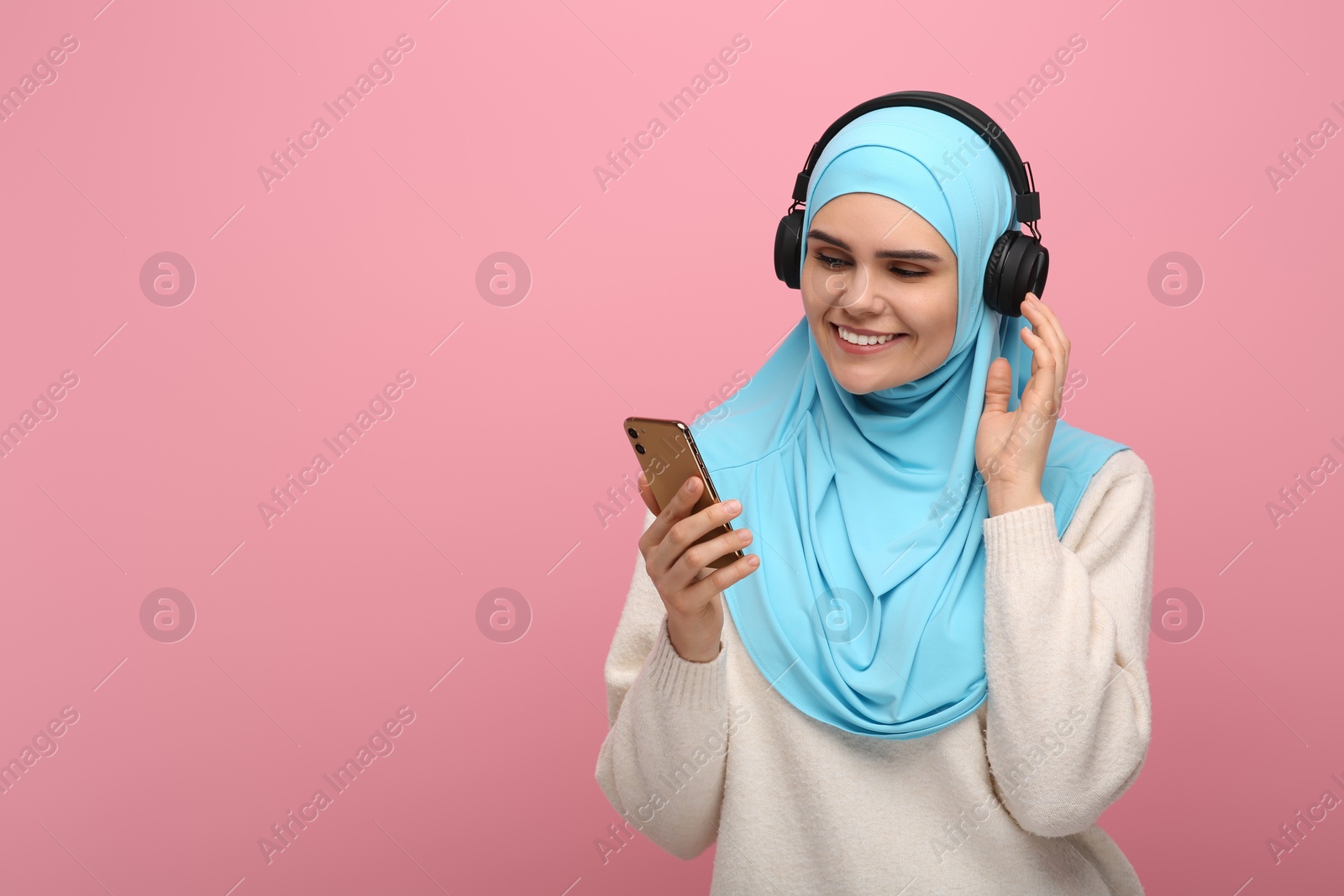 Photo of Muslim woman in hijab and headphones listening to music with smartphone on pink background, space for text