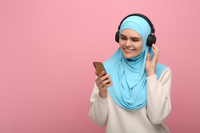 Photo of Muslim woman in hijab and headphones listening to music with smartphone on pink background, space for text