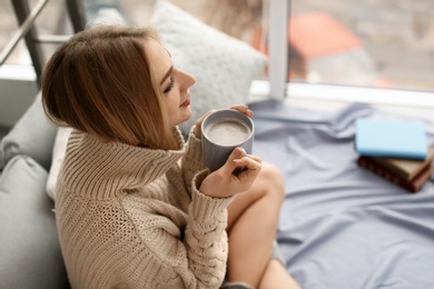 Beautiful young woman in knitted sweater sitting with cup of coffee at home. Winter atmosphere