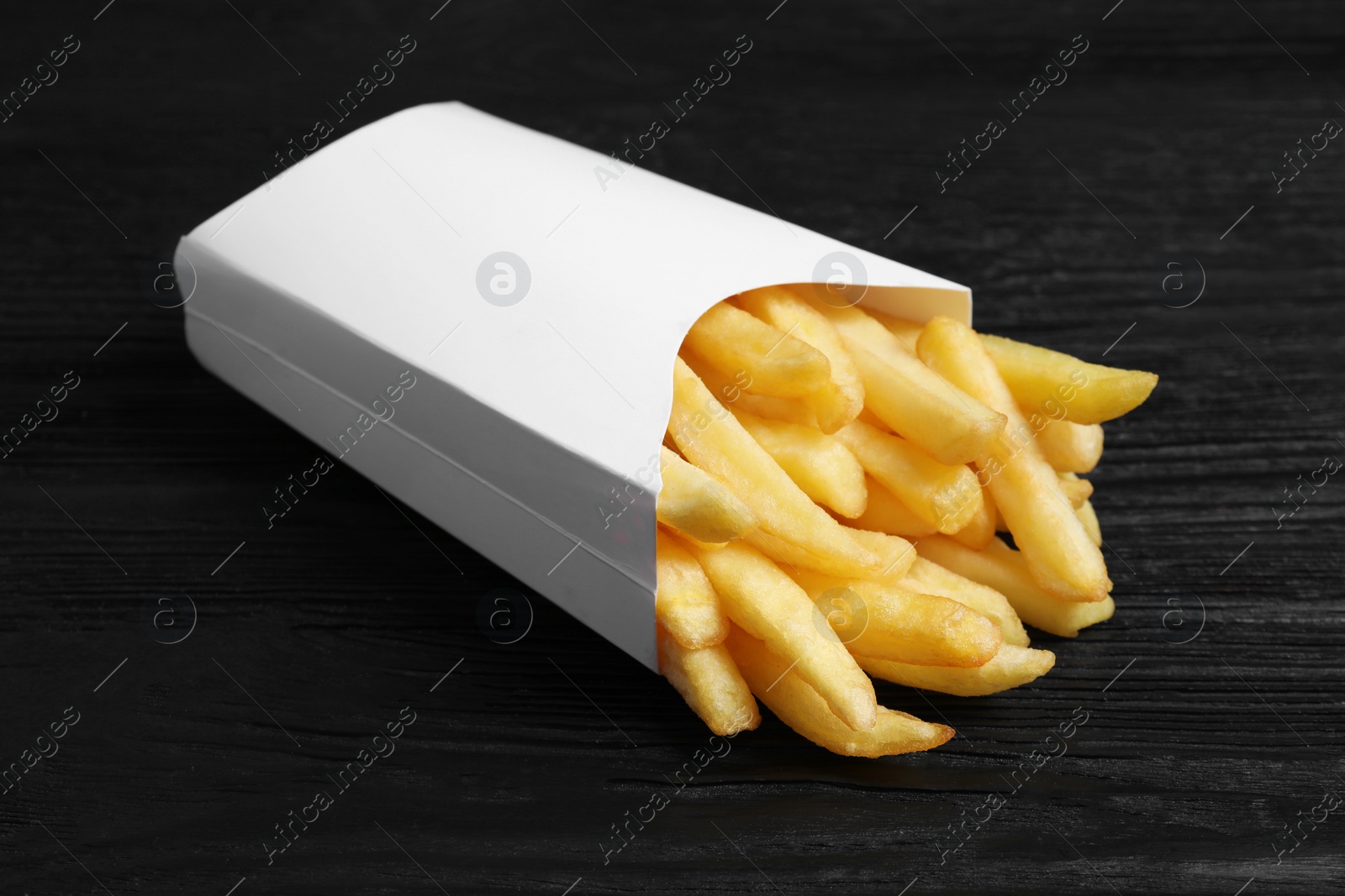 Photo of Delicious french fries in paper box on black wooden table, closeup