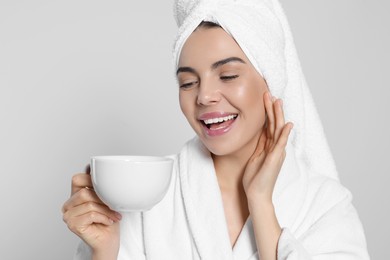 Young woman in bathrobe with towel and cup of drink on light grey background. Spa treatment