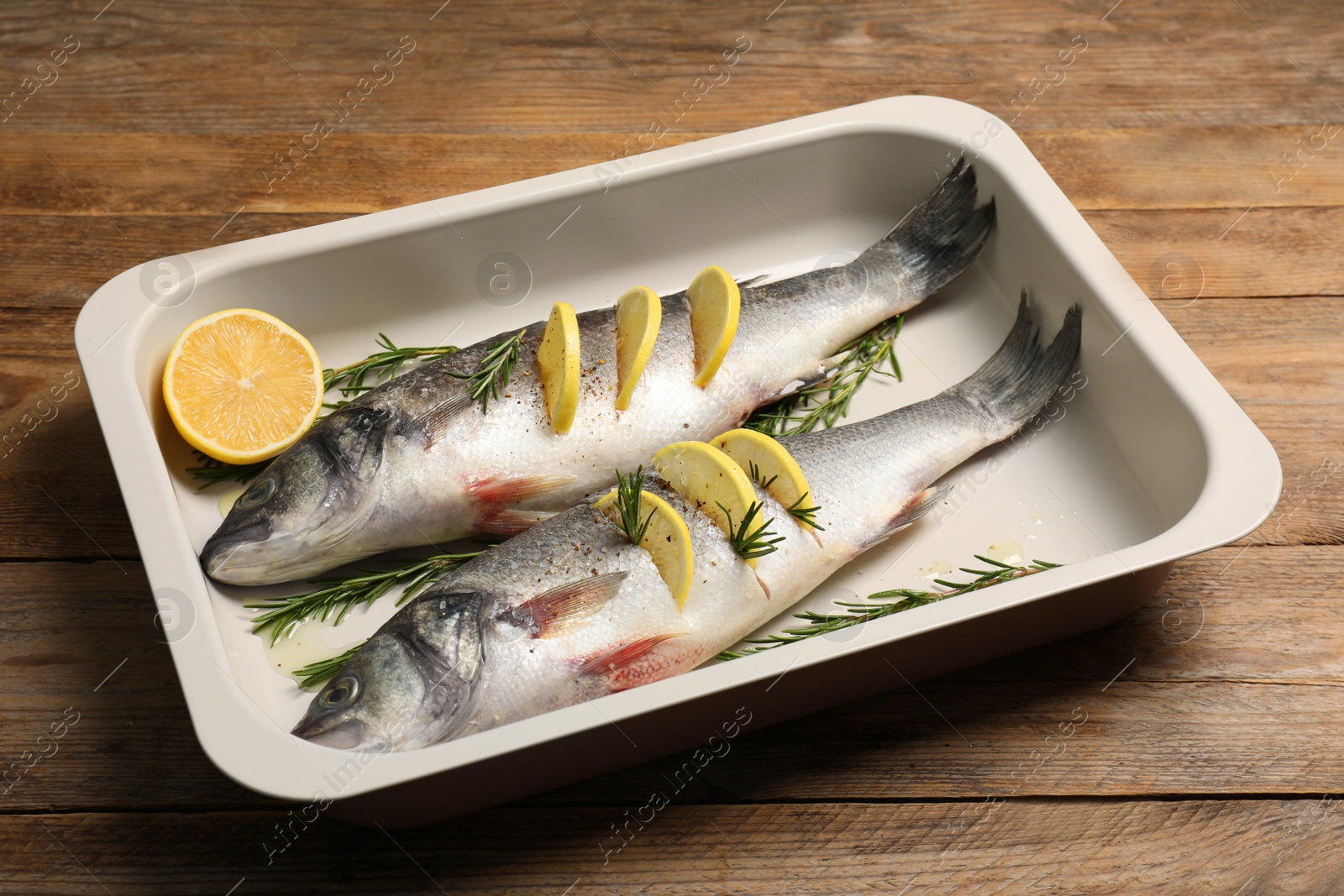 Photo of Baking tray with raw sea bass fish, lemon and rosemary on wooden table