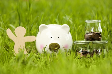 Photo of Cute piggy bank, wooden person and jars with coins on green grass in park