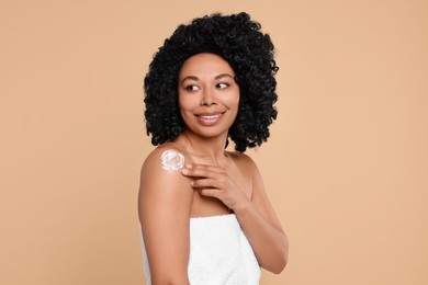 Young woman applying body cream onto shoulder on beige background