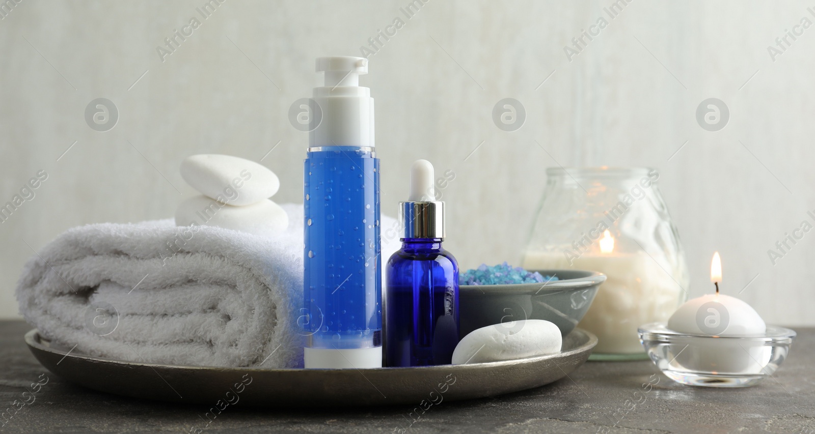 Photo of Spa composition. Cosmetic products, terry towel, burning candles and sea salt on gray table against light background