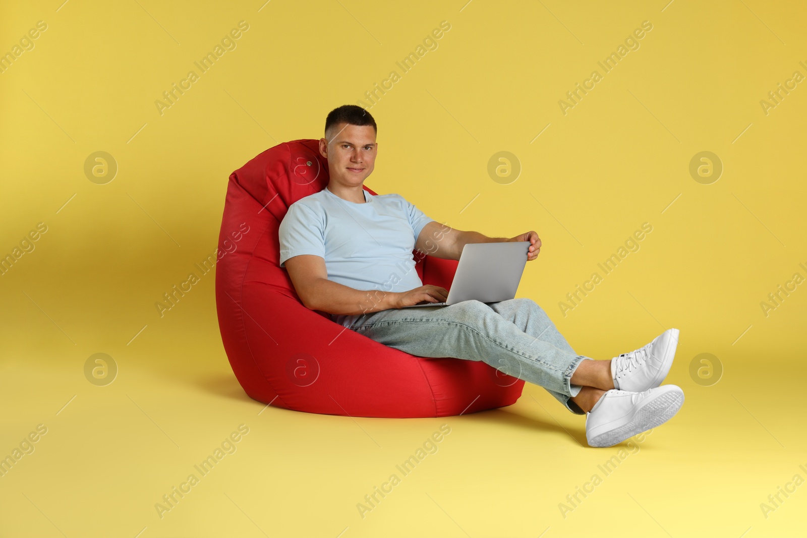 Photo of Handsome man with laptop on red bean bag chair against yellow background