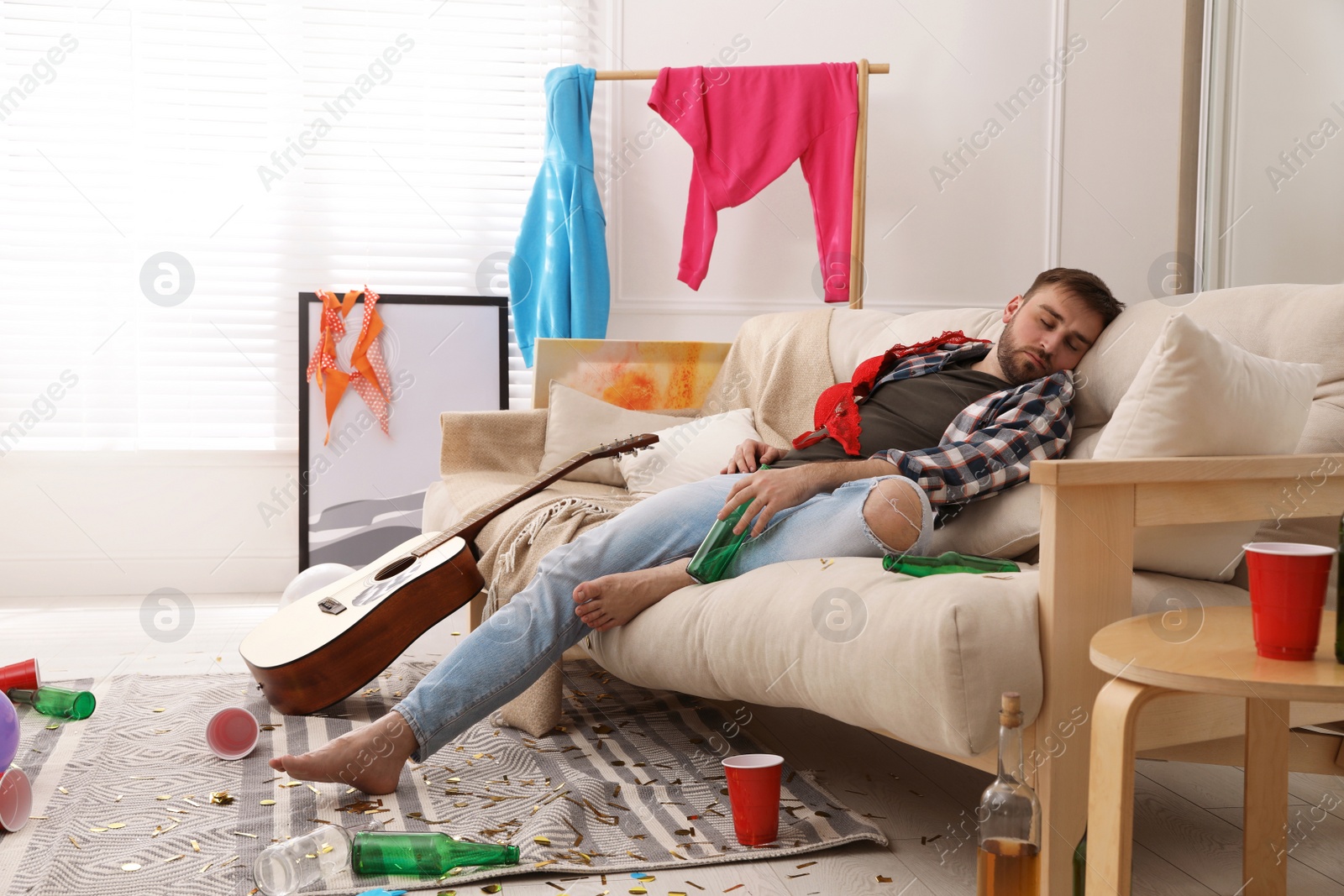 Photo of Young man with bottle of beer sleeping on sofa in messy room after party