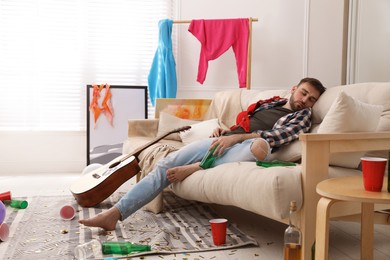 Photo of Young man with bottle of beer sleeping on sofa in messy room after party