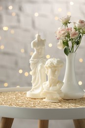 Photo of Beautiful David bust and Venus De Milo candles near flowers on table against blurred lights
