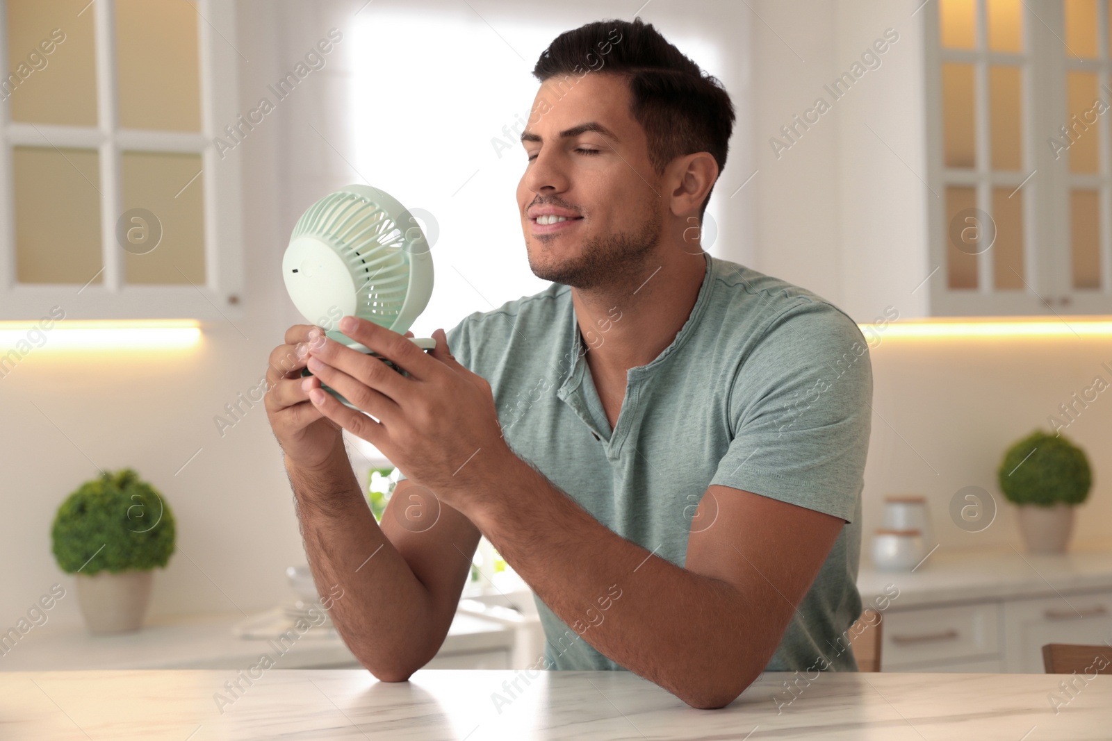 Photo of Man enjoying air flow from portable fan at table in kitchen. Summer heat