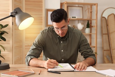 Photo of Man drawing in sketchbook with pencil at wooden table indoors