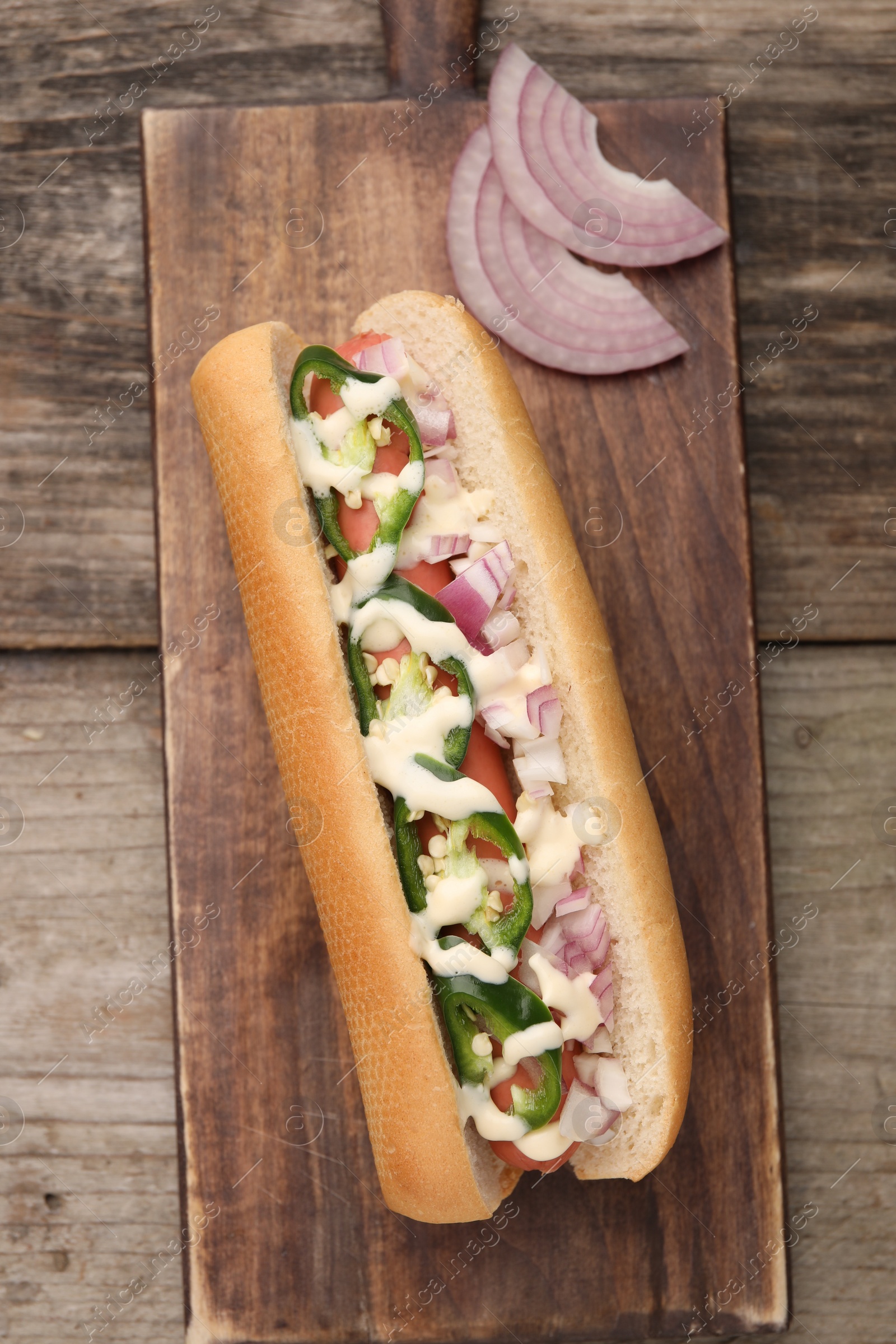 Photo of Delicious hot dog with onion, chili pepper and sauce on wooden table, top view