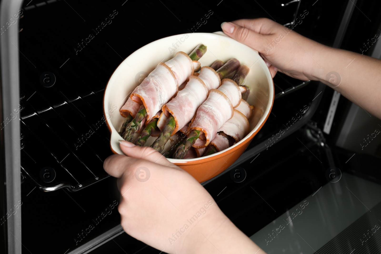 Photo of Woman putting ceramic baking dish with bacon wrapped asparagus in oven, closeup