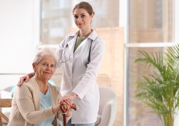 Photo of Nurse assisting elderly woman with cane indoors. Space for text