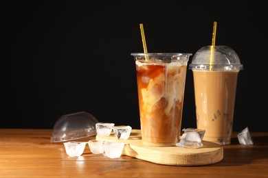 Photo of Refreshing iced coffee with milk in takeaway cups on wooden table against black background, space for text