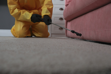 Photo of Pest control worker in protective suit spraying insecticide under sofa indoors, closeup