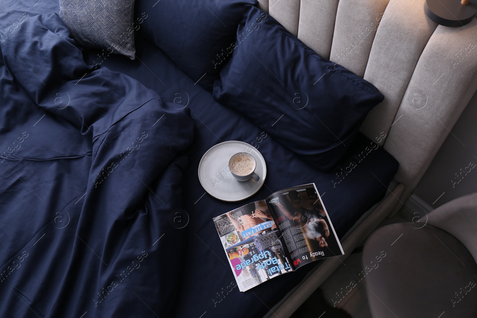 Photo of Open magazine and cup of coffee on bed with stylish silky linens in room, above view