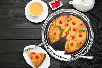 Photo of Delicious cut pineapple pie with cherry and tea served on black wooden table, flat lay