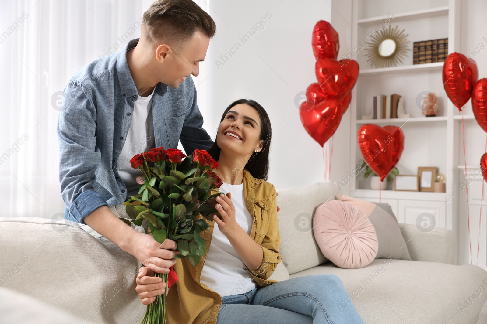 Photo of Boyfriend presenting beautiful bouquet of roses to his girlfriend in room decorated with heart shaped balloons. Valentine's day celebration