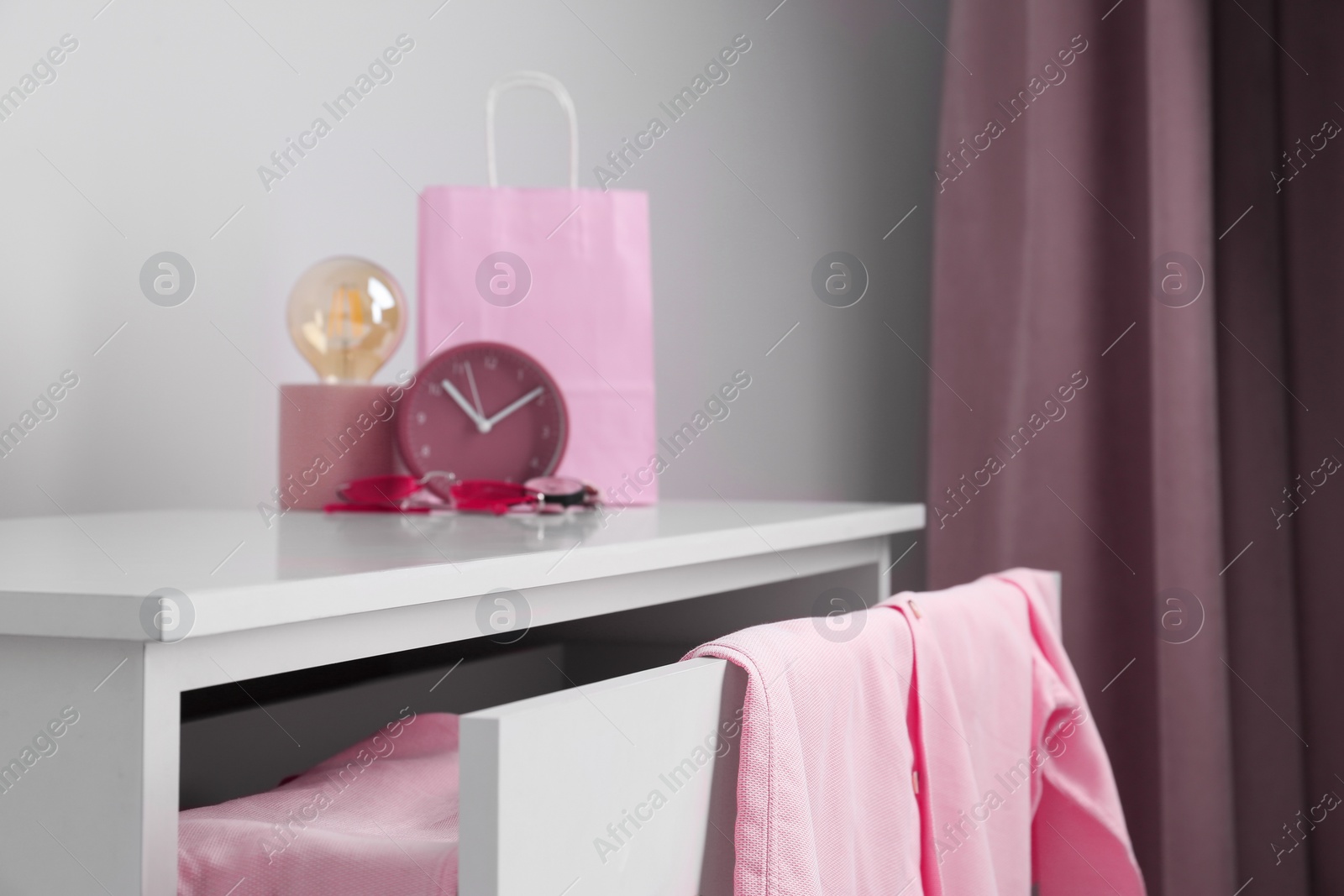 Photo of White chest of drawers with pink clothes, clock, sunglasses and decor indoors