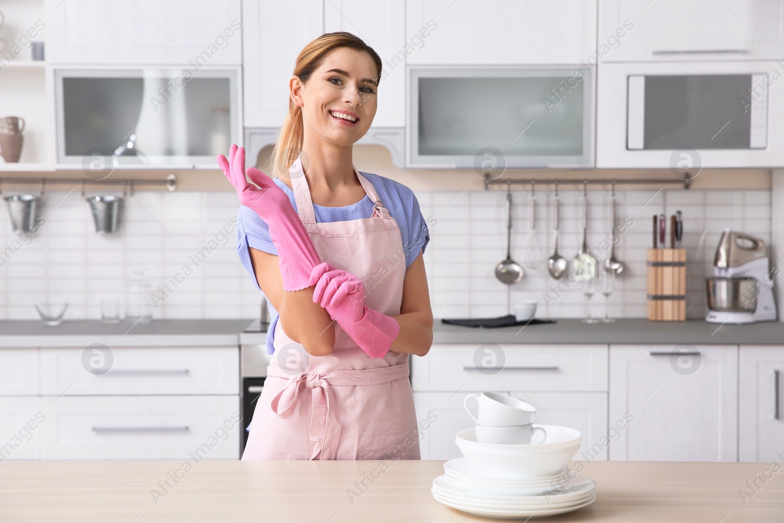 Photo of Woman wearing rubber gloves near table with clean dishes and cups in kitchen