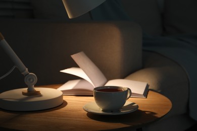 Photo of Stylish lamp, book and cup of tea on side table near sofa indoors