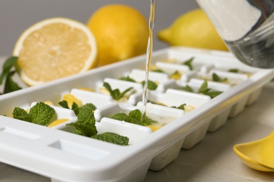 Pouring water into ice cube tray with mint and lemon on table, closeup