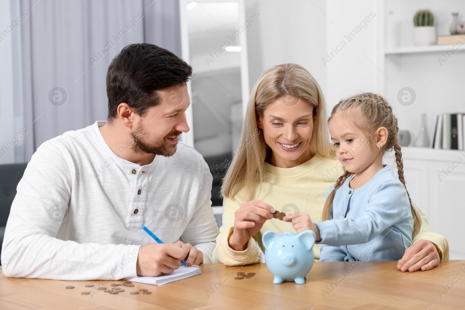 Photo of Planning budget together. Little girl with her family putting coins into piggybank at table indoors