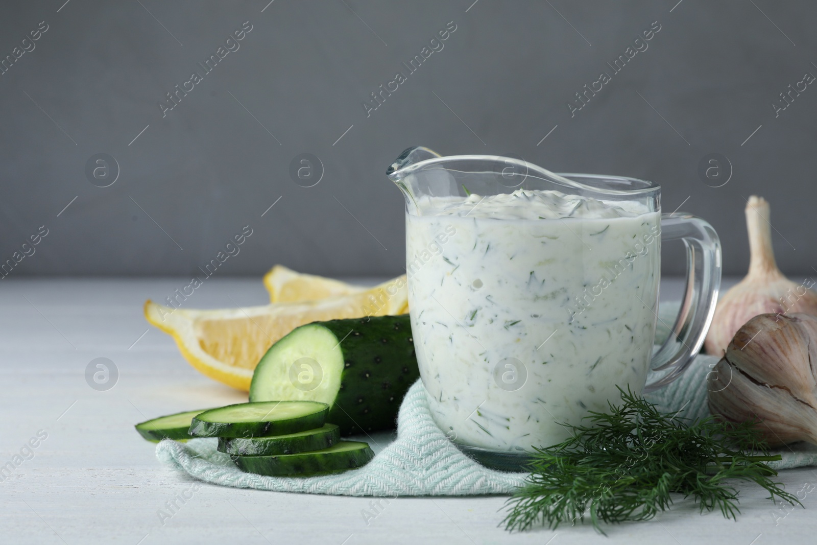 Photo of Glass jug of Tzatziki cucumber sauce with ingredients on grey background, space for text