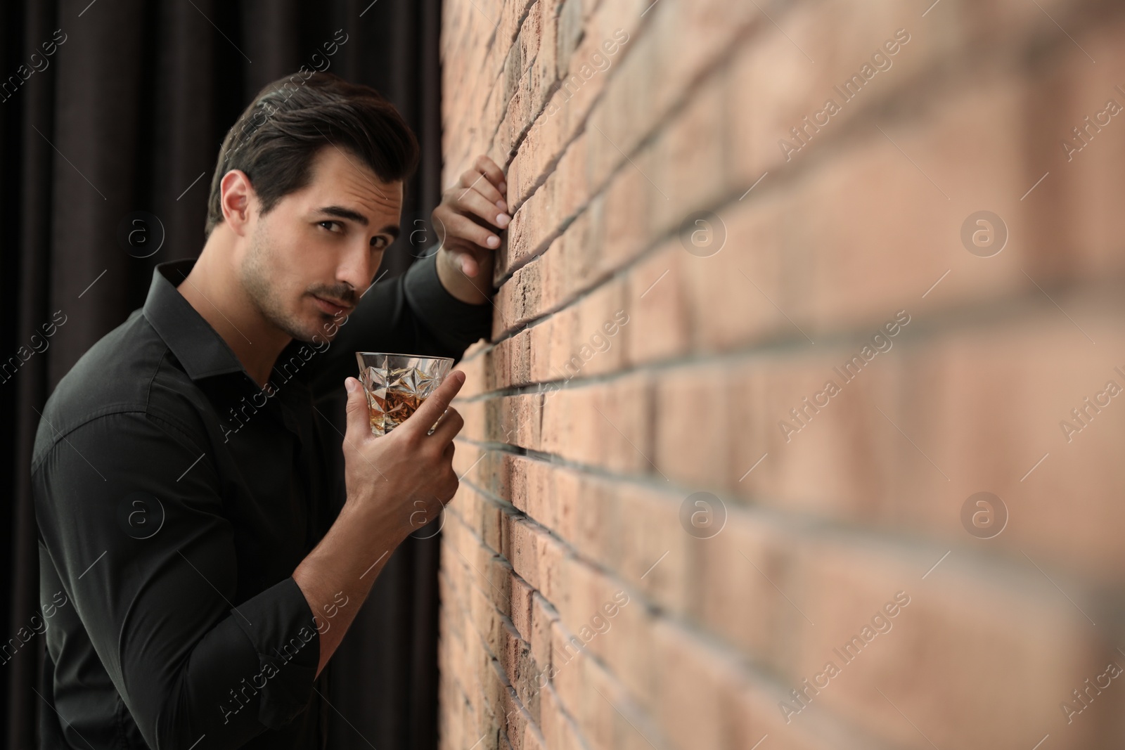 Photo of Young man with glass of whiskey near brick wall indoors. Space for text